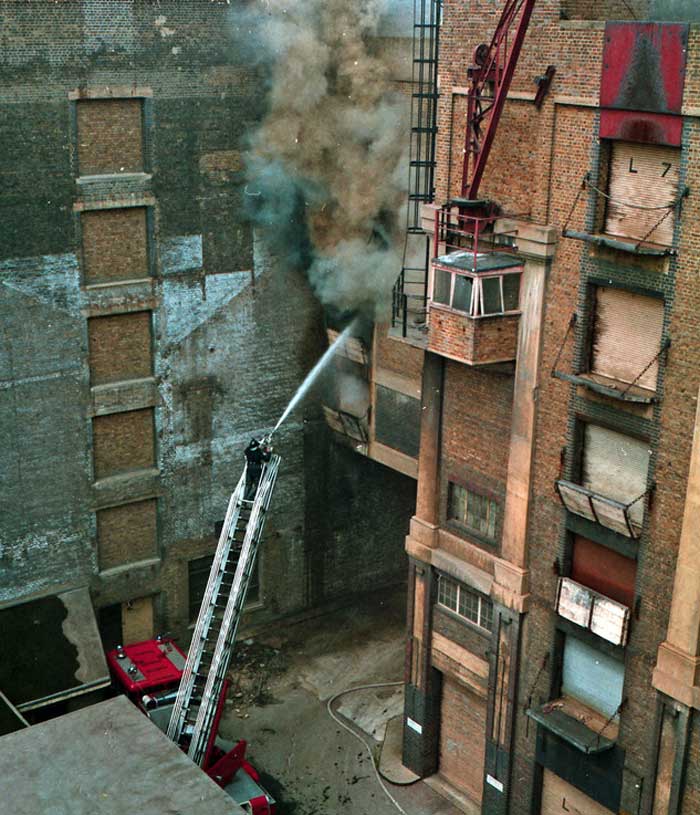 Wider angle shot of Wilson's Wharf building and a firefighter putting the fire out using a hose 