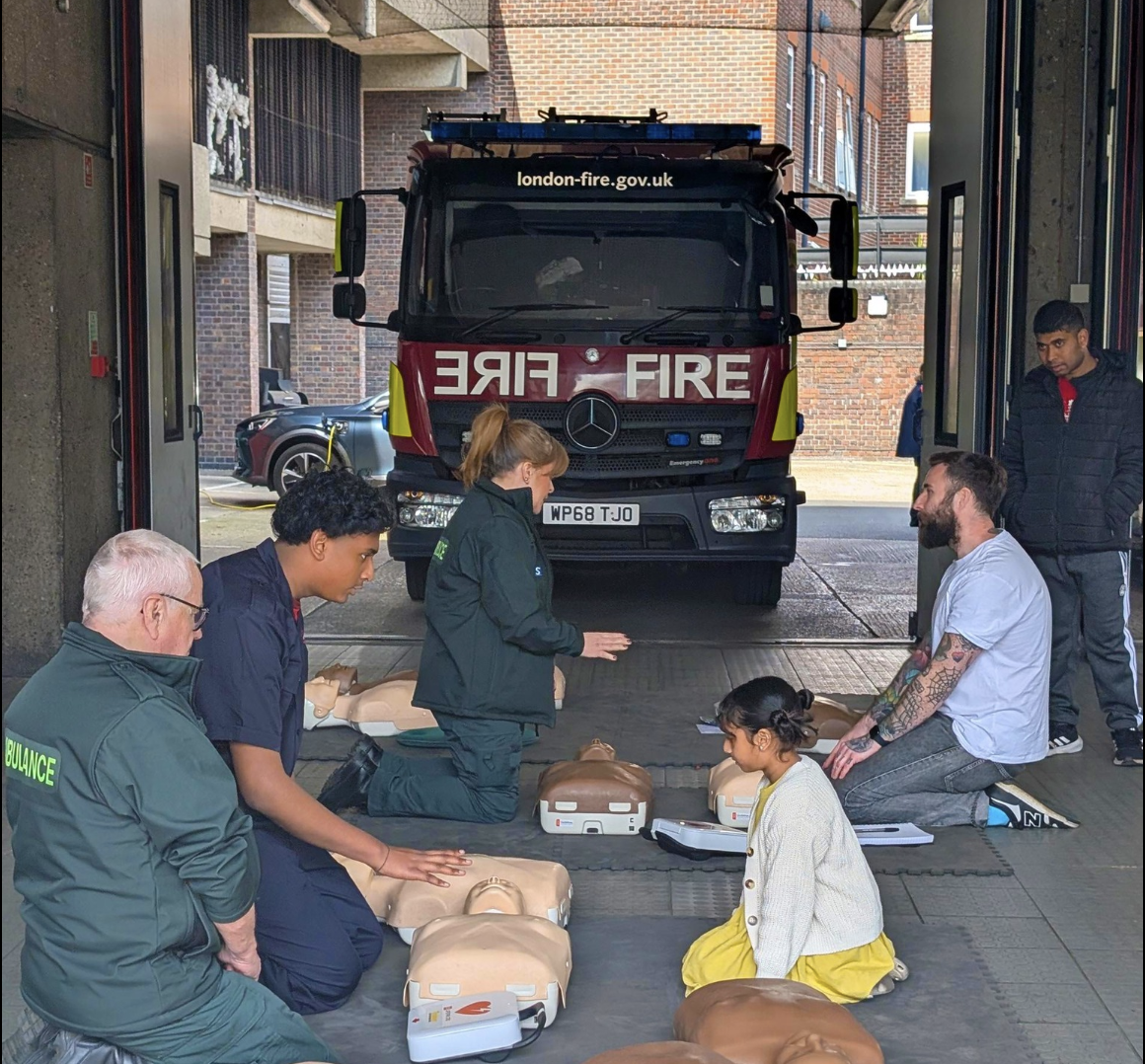 A group of people, including emergency services personnel and civilians, are participating in a CPR training session inside a fire station. Mannequins are laid out on mats for practice. A fire truck marked “london-fire.gov.uk” is parked behind them inside the station. A young girl in a yellow dress is also present, observing the training.