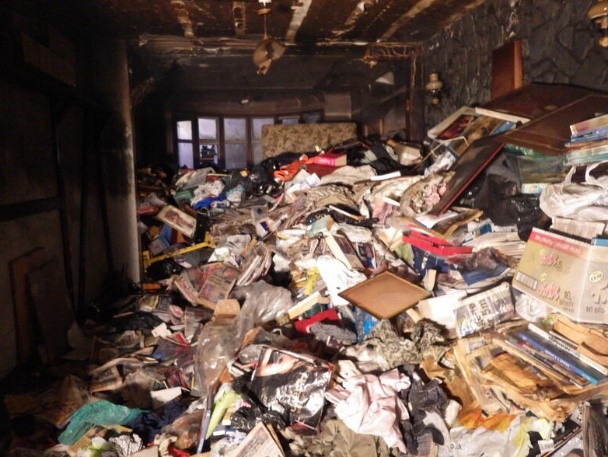 A fire damaged room with lots of possessions piled high, including books and clothes