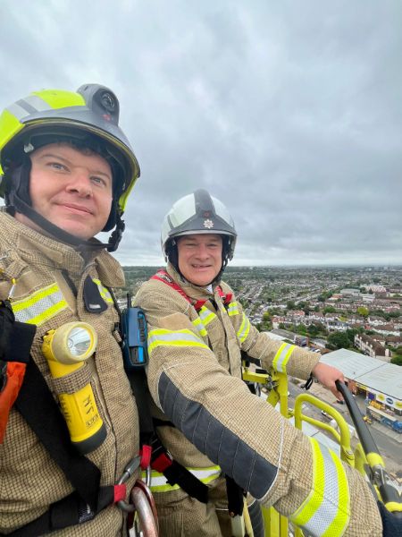 A firefighter on the left and Sir Ed Davey MP on the right in the cage of the 64-metre turntable ladder wearing full firefighting gear.
