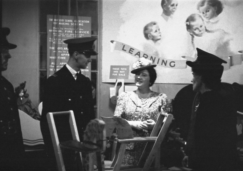 Black-and-white photograph of an indoor exhibition featuring handmade toys and furniture. A lady in suit, necklace and hat stands between uniformed men and women around a wooden rocking horse, examining or discussing the display. Behind them is a large poster showing illustrations of children and the word “LEARNING” in large letters. 
