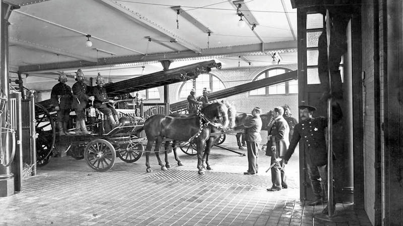 Firefighters are preparing the horses and fire engines to leave Kensington Fire Station in around 1905. Two firefighters have used the sliding pole to reach the appliance bay.