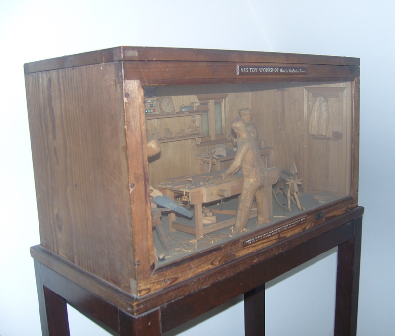 Wooden display case on a stand containing a miniature diorama of a toy workshop. Inside the case, a carved figure stands at a workbench surrounded by woodworking tools, shelves, a stool, and wood shavings on the floor. The interior is finished to resemble a workshop with wooden walls and furnishings. A label at the top of the case reads “NFS TOY WORKSHOP.” The display is photographed against a plain light-coloured background.