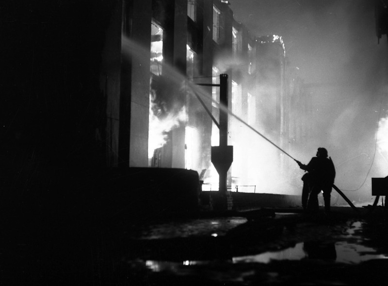 A Blitz bombing raid on Pimlico, London in 1941. Firefighters are tackling a fire in a nearby building, spraying water from hoses. They are silhouetted against the bright light of the fire in the background.