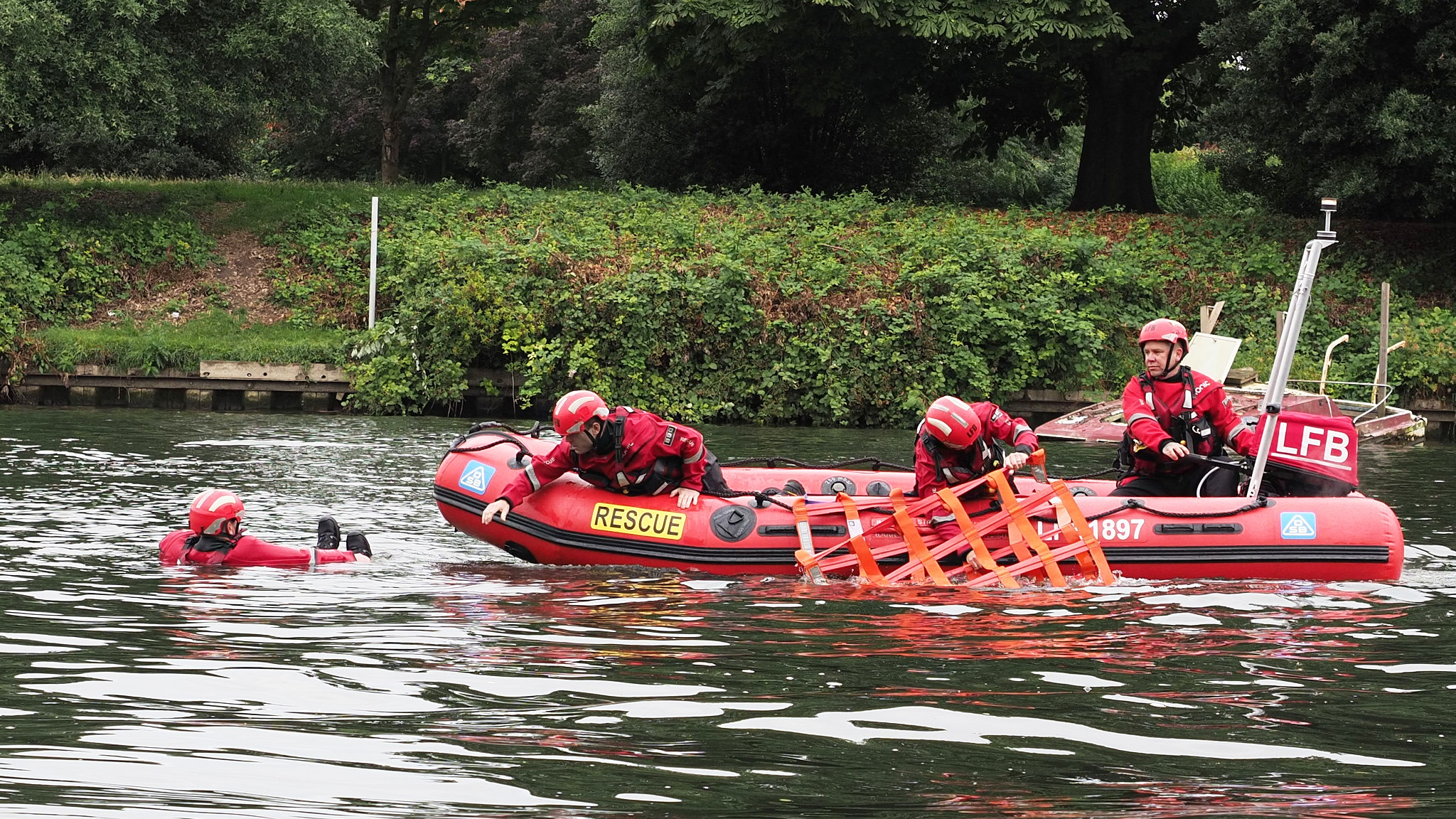 Three firefighters in lifejackets sitting in an inflatable rescue boat, working to rescue another firefighter wearing a lifejacket floating in the water as part of a training exercise.