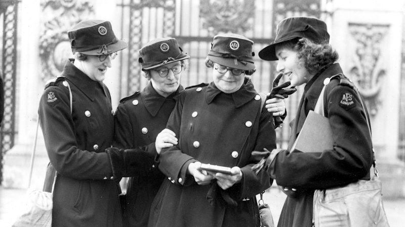 A black‑and‑white photo of four uniformed people standing together outdoors, looking at a small item one of them is holding.
