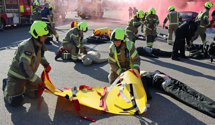 Two firefighters holding a yellow stretcher. Lots of other firefighters are training with dummy patients in the background. 