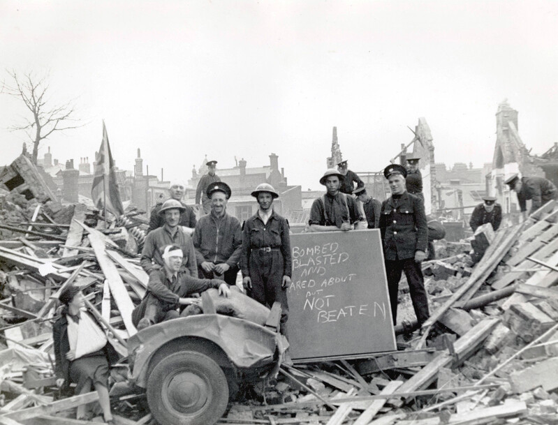 Black-and-white photograph showing a group of men in uniform and helmets and local residents standing among the rubble of bomb-damaged buildings. In the foreground, a damaged vehicle is surrounded by debris and broken timber. Several people pose beside a handwritten sign that can be partially read, “Bombed, Blasted and ?ared About But Not Beaten.” Partially standing walls and ruined houses are visible in the background, along with a British flag among the wreckage.