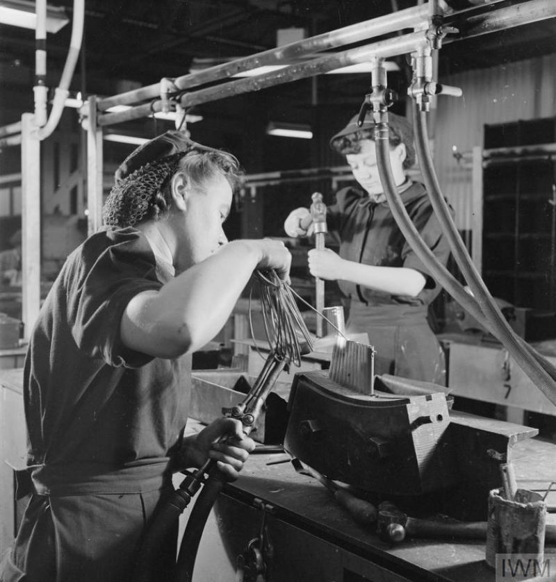 Black-and-white photograph of two factory workers operating industrial equipment at a production workstation. One worker in the foreground holds a hose connected to machinery while working on a metal mould or component. A second worker stands behind, adjusting a control or tool. Overhead pipes and hoses run across the workspace, and industrial machinery surrounds the workbench