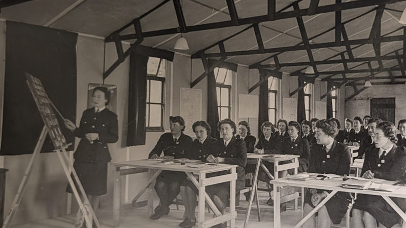 A classroom scene showing a group of uniformed people seated at desks while another person stands at the front beside a board, appearing to lead a training session.