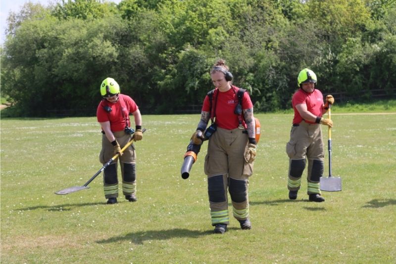 Three firefighters on a field of grass demonstrating a backpack blower and wildfire beaters.
