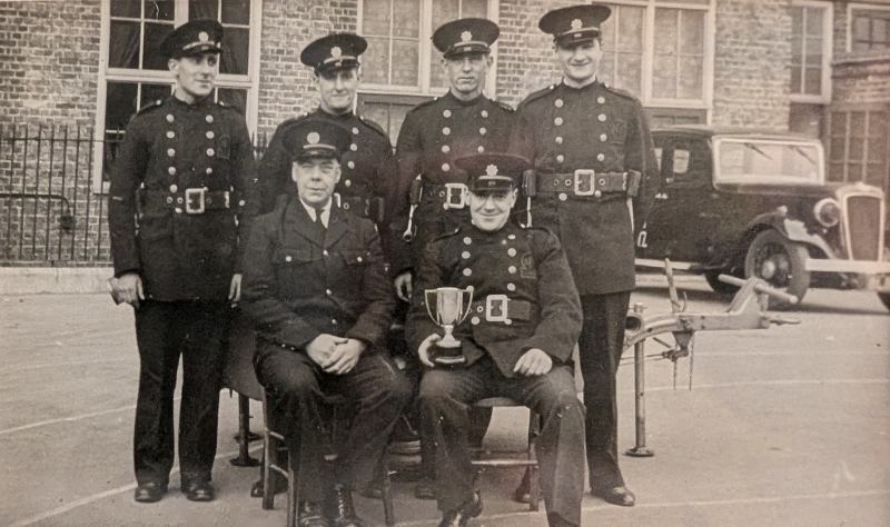 Photograph of Auxiliary Fire Service firefighters at a Fulham sub-station during the Second World War, showing a station team gathered for a group portrait with an award trophy, trailer pump and service vehicle.