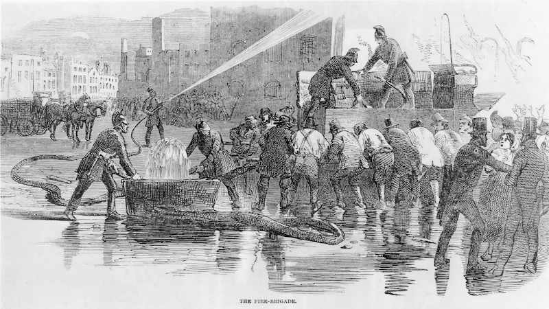 With water pooling on the ground, many volunteers gather around the fire engine to manually pump the sides of the engine to push the water through the hose towards the fire, directed by a firefighter from the London Fire Engine Establishment
