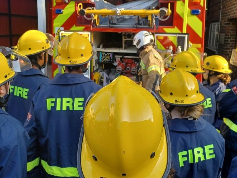 Fire cadets in yellow helmets listening to a firefighter.