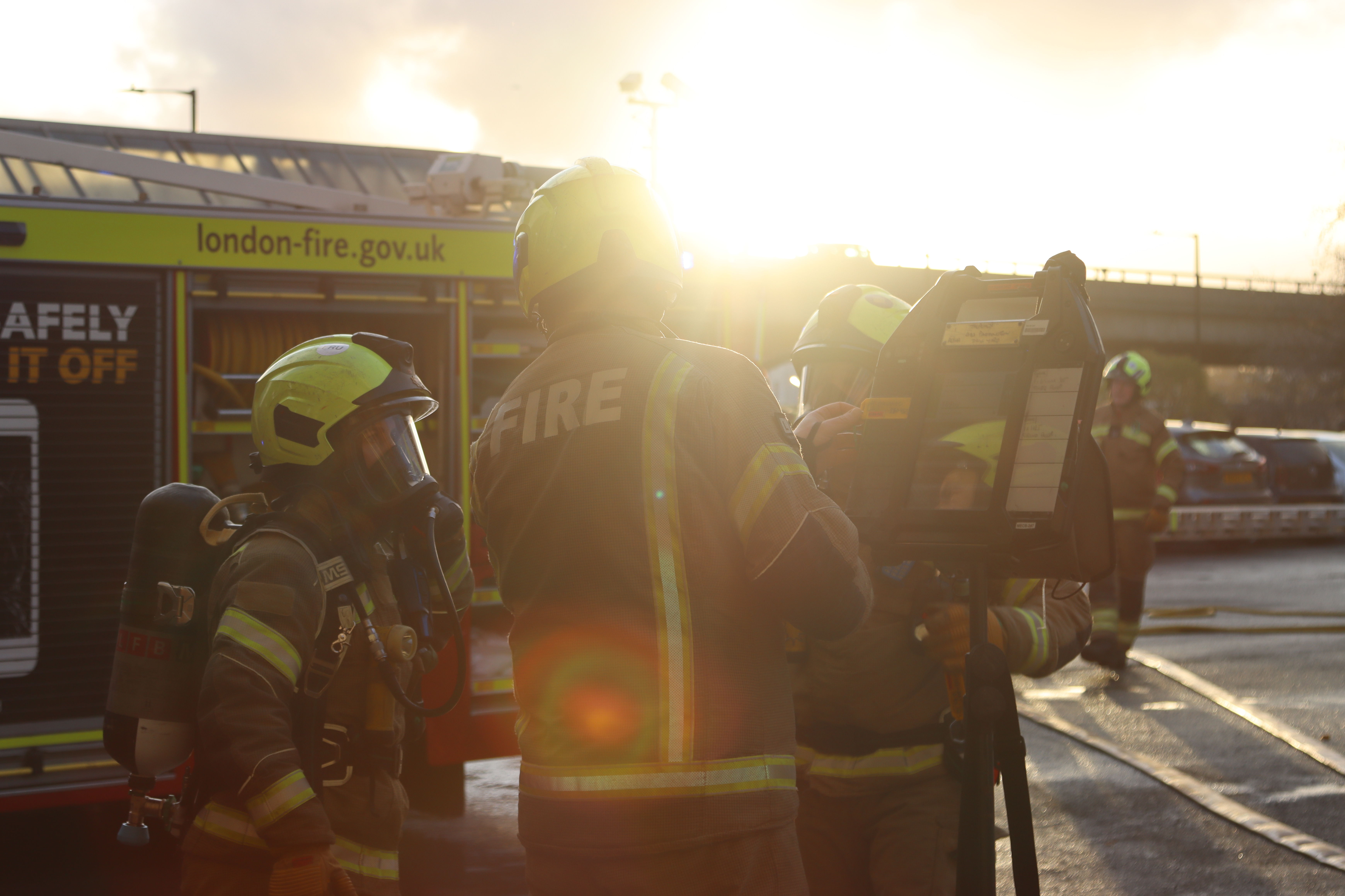 Three firefighters stand next to a breathing apparatus board with breathing apparatus on and a fire engine in the background. It is sunset and another firefighter is in the background walking towards them.