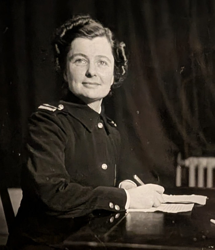 Photograph of a uniformed person seated at a desk, holding a pen and looking slightly upward, with papers in front of them and a dark curtain behind.