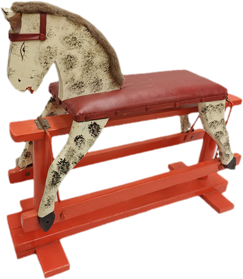 Photograph of a painted wooden rocking horse. The horse has a cream-coloured body with black mottled markings, a grey fabric mane, and a red bridle. A long red padded seat forms the horse's back, allowing it to be used as a bench. The horse stands on a sturdy red wooden rocking frame with cross supports. The object is shown against a plain white background, highlighting its decorative paintwork.