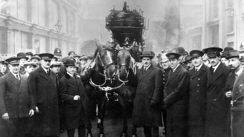 A large group of men in London Fire Brigade and Police uniforms surround two horses, tacked up to pull a turntable fire ladder in 1921.