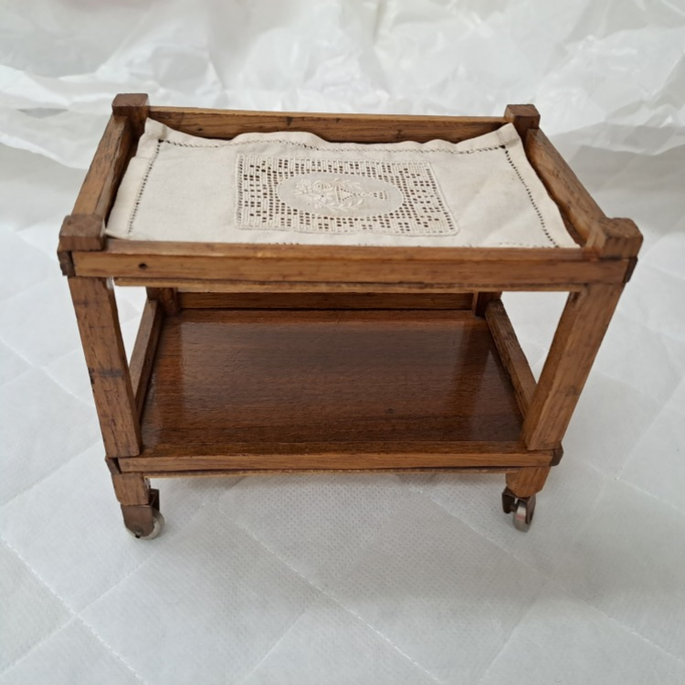 Small wooden two-tier trolley on four caster wheels. The top tray is lined with a white cloth featuring a decorative lace insert, while the lower shelf is polished wood. The trolley is photographed against a plain white fabric background, showing its simple rectangular frame and vintage-style construction.